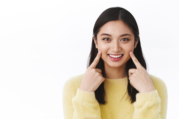 Atractiva mujer sonriente con cabello corto oscuro apuntando a las mejillas y sonriendo, recomienda usar productos para el cuidado de la piel o cosméticos para la piel, de pie en la pared blanca