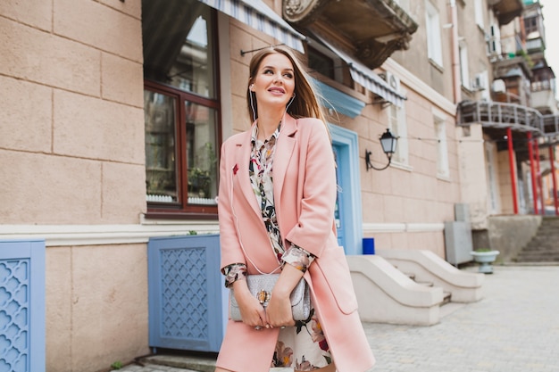 Atractiva mujer sonriente elegante caminando por las calles de la ciudad en abrigo rosa, escuchando música en auriculares