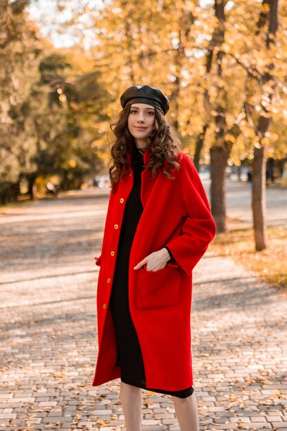 Atractiva mujer sonriente con estilo con el pelo rizado caminando en el parque vestida con abrigo rojo cálido otoño moda moda, estilo callejero, con sombrero de boina