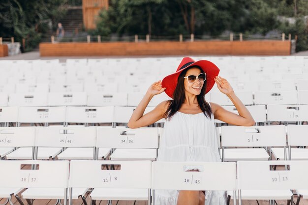 Atractiva mujer sonriente feliz vestida con vestido blanco, sombrero rojo, gafas de sol sentado en el teatro al aire libre de verano en silla sola, tendencia de moda de estilo callejero de primavera