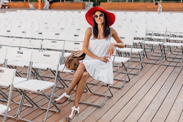 Atractiva mujer sonriente feliz vestida con vestido blanco, sombrero rojo, gafas de sol sentado en el teatro al aire libre de verano solo, muchas sillas, tendencia de moda de estilo callejero de primavera, saludando con la mano hola