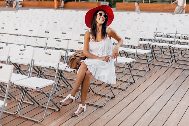 Atractiva mujer sonriente feliz vestida con vestido blanco, sombrero rojo, gafas de sol sentado en el teatro al aire libre de verano solo, muchas sillas, tendencia de moda de estilo callejero de primavera, saludando con la mano hola