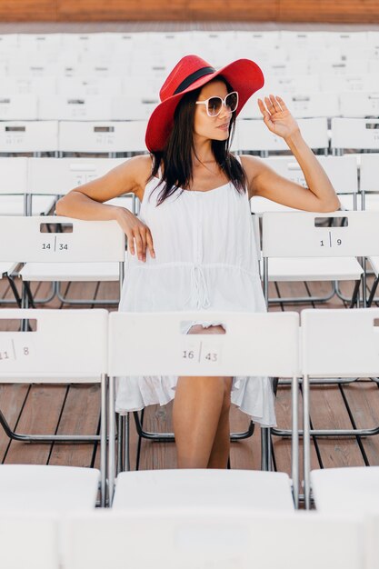 Atractiva mujer sonriente feliz vestida con vestido blanco, sombrero rojo, gafas de sol sentado en el teatro al aire libre de verano solo, muchas sillas, tendencia de moda de estilo callejero de primavera