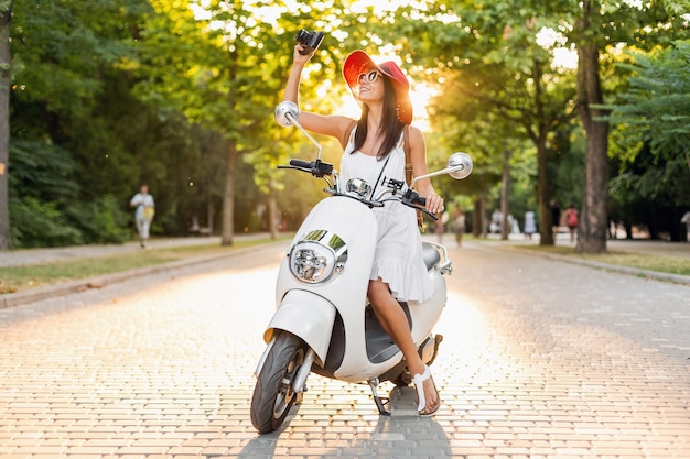 Foto gratuita atractiva mujer sonriente montando en moto en la calle en traje de estilo veraniego con vestido blanco y sombrero rojo viajando de vacaciones, tomando fotografías en la cámara de fotos vintage