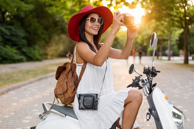 Atractiva mujer sonriente montando en moto en la calle en traje de estilo veraniego con vestido blanco y sombrero rojo viajando de vacaciones, tomando fotografías en la cámara de fotos vintage