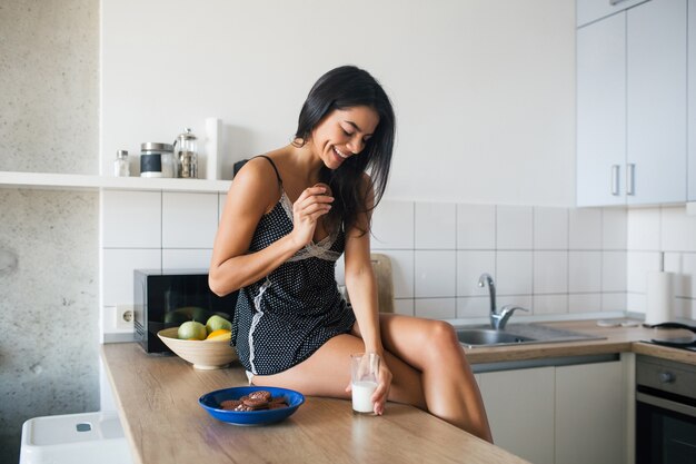 Atractiva mujer sonriente en pijama desayunando en la cocina por la mañana, comiendo galletas y bebiendo leche, estilo de vida saludable