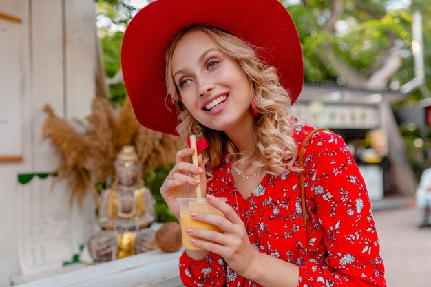 Atractiva mujer sonriente rubia elegante con sombrero rojo de paja y blusa traje de moda de verano bebiendo batido de cóctel de frutas naturales