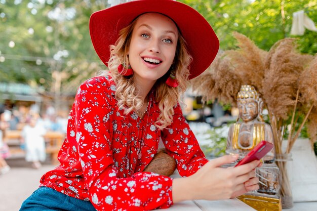 Atractiva mujer sonriente rubia elegante con sombrero rojo de paja y blusa traje de moda de verano sosteniendo usando café de teléfono inteligente