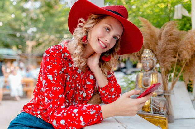 Atractiva mujer sonriente rubia elegante con sombrero rojo de paja y blusa traje de moda de verano sosteniendo usando café de teléfono inteligente