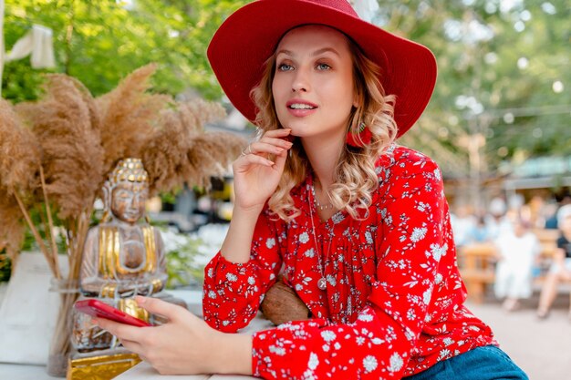 Atractiva mujer sonriente rubia elegante con sombrero rojo de paja y blusa traje de moda de verano sosteniendo usando café de teléfono inteligente