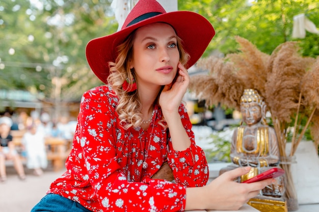 Atractiva mujer sonriente rubia elegante con sombrero rojo de paja y blusa traje de moda de verano sosteniendo usando café de teléfono inteligente