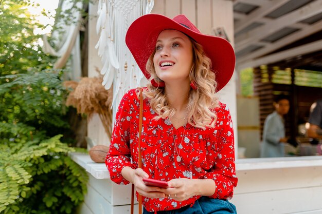 Atractiva mujer sonriente rubia elegante con sombrero rojo de paja y blusa traje de moda de verano sosteniendo usando café de teléfono inteligente