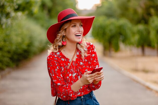 Atractiva mujer sonriente rubia elegante en traje de moda de verano de sombrero rojo de paja y blusa con teléfono