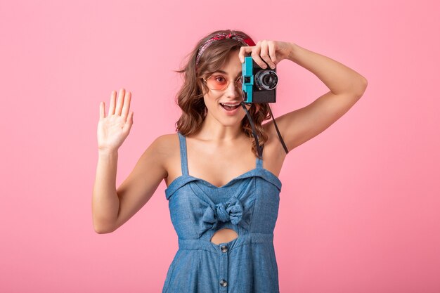 Atractiva mujer sonriente tomando una foto con una cámara vintage, apuntando con el dedo hacia arriba, vestido con un vestido de mezclilla y gafas de sol aisladas sobre fondo rosa