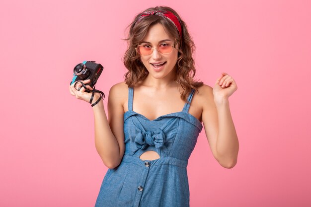 Atractiva mujer sonriente tomando una foto con una cámara vintage, apuntando con el dedo hacia arriba, vestido con un vestido de mezclilla y gafas de sol aisladas sobre fondo rosa