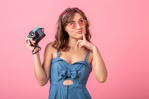 Atractiva mujer sonriente tomando una foto con una cámara vintage, apuntando con el dedo hacia arriba, vestido con un vestido de mezclilla y gafas de sol aisladas sobre fondo rosa