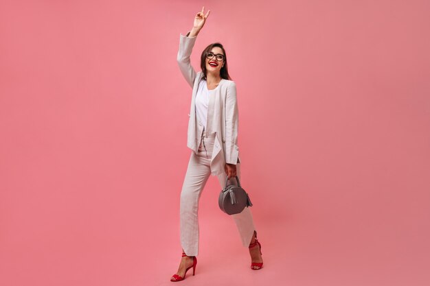 Atractiva mujer sonriente en traje blanco moderno y anteojos sostiene el bolso y muestra el signo de la paz sobre fondo rosa aislado.