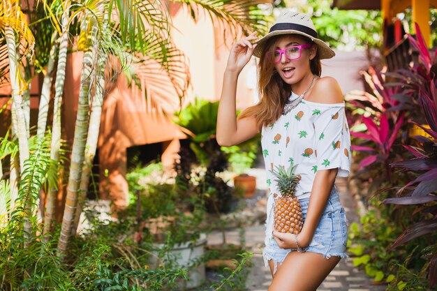 Atractiva mujer sonriente de vacaciones en camiseta impresa sombrero de paja moda de verano, manos sosteniendo piña