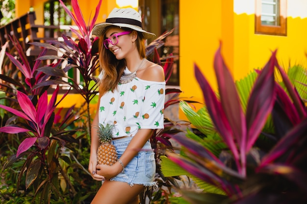 Atractiva mujer sonriente de vacaciones en camiseta impresa sombrero de paja moda de verano, manos sosteniendo piña