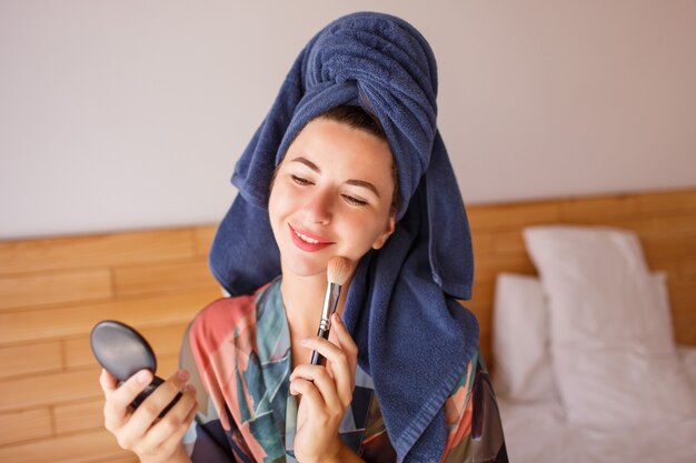 Atractiva mujer vestida con bata de baño, toalla de ducha en la cabeza se prepara para el maquillaje, cepillo en mano
