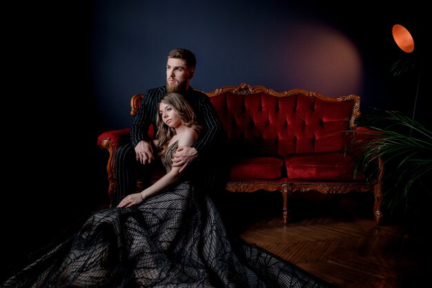 Atractiva mujer vestida con elegante vestido de noche y guapo con barba vestido con traje negro tomados de la mano y sentado en el lujoso sofá rojo