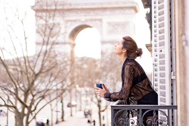 Atractiva mujer yang en pijama está tomando café en el balcón por la mañana en la ciudad de París. vista del arco triunfal.