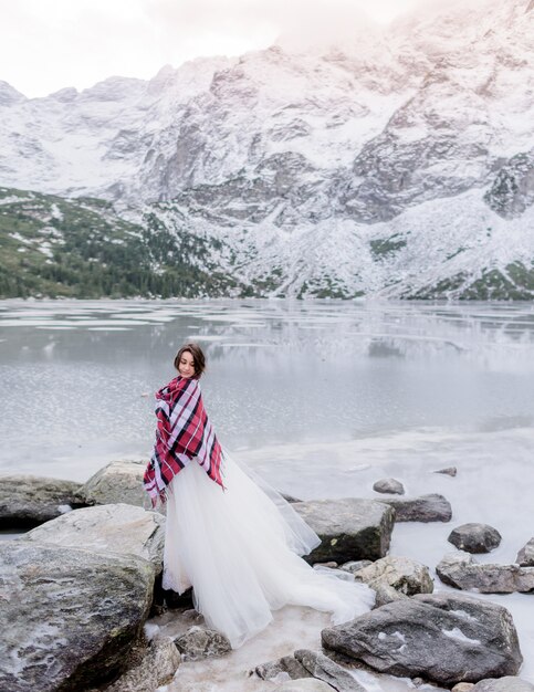 Atractiva novia cubierta con una manta está de pie sobre la roca cerca del lago congelado rodeado de montañas