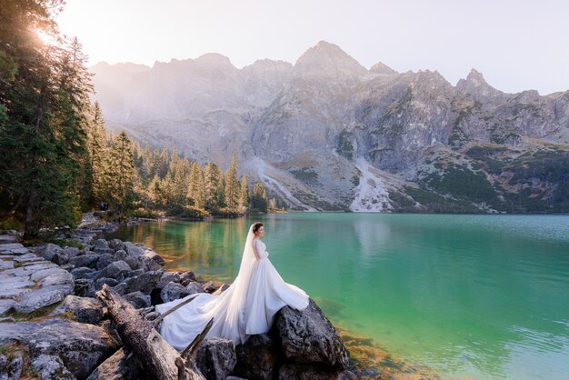 Atractiva novia está de pie cerca del lago Highland con pintorescas vistas de las montañas de otoño