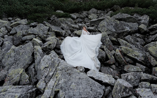 Atractiva novia está de pie sobre las enormes rocas grises vestidas con un vestido ceremonial blanco