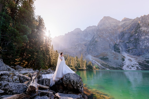 Atractiva novia está de pie sobre la roca con una vista impresionante del lago Highland con agua de color verde en el día soleado, las montañas Tatry