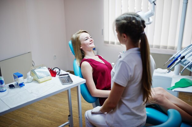 Atractiva paciente vestida de violeta rojiza tendida en el sillón dental mientras una dentista trata sus dientes con instrumentos especiales