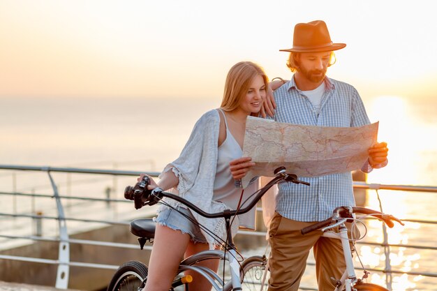 Atractiva pareja feliz de amigos que viajan en verano en bicicleta, hombre y mujer con cabello rubio boho hipster estilo moda divirtiéndose juntos