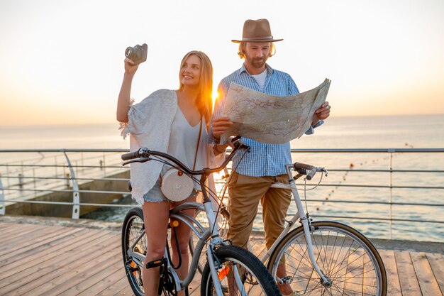Atractiva pareja feliz viajando en verano en bicicleta, hombre y mujer con cabello rubio boho estilo hipster divirtiéndose juntos, mirando en el mapa de turismo tomando fotos en la cámara