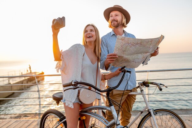 Atractiva pareja feliz viajando en verano en bicicleta, hombre y mujer con cabello rubio boho estilo hipster divirtiéndose juntos, mirando en el mapa de turismo tomando fotos en la cámara