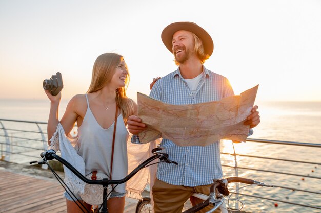 Atractiva pareja feliz viajando en verano en bicicleta, hombre y mujer con cabello rubio boho estilo hipster divirtiéndose juntos, mirando en el mapa de turismo tomando fotos en la cámara