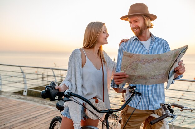 Atractiva pareja feliz viajando en verano en bicicleta, hombre y mujer con cabello rubio boho hipster estilo moda divirtiéndose juntos, mirando en el mapa de turismo