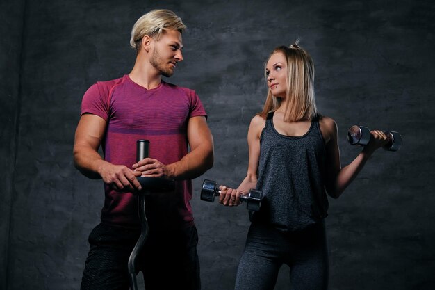 Una atractiva pareja de fitness de hombres deportivos sostiene pesas y una mujer rubia delgada sostiene pesas sobre fondo gris