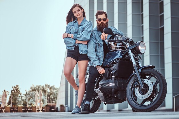 Atractiva pareja hipster - hombre brutal barbudo con gafas de sol y chaqueta de jeans sentado en una motocicleta retro y su joven chica sensual parada cerca, posando contra rascacielos.