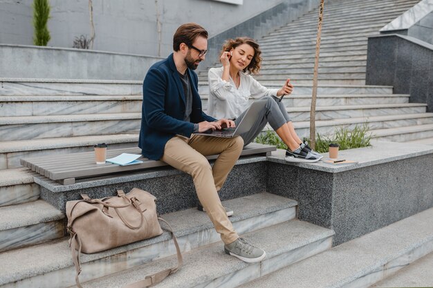 Atractiva pareja de hombre y mujer sentados en las escaleras en el centro urbano de la ciudad, trabajando juntos en la computadora portátil