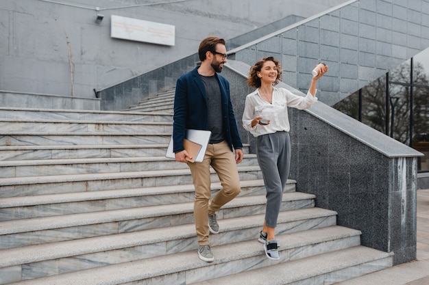 Atractiva pareja de hombre y mujer subiendo escaleras en el centro urbano de la ciudad, sosteniendo portátil, discutiendo