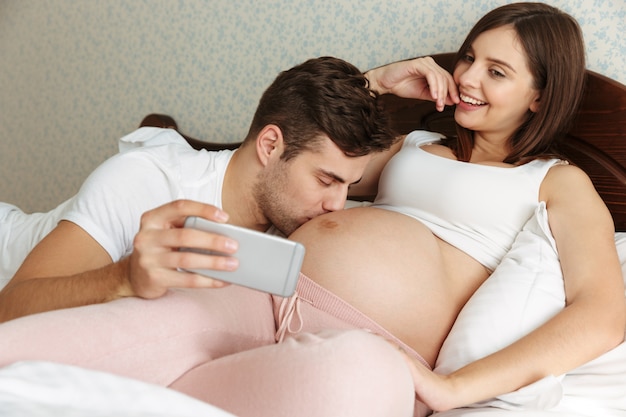 Atractiva pareja joven embarazada tomando selfie mientras está acostado en la cama