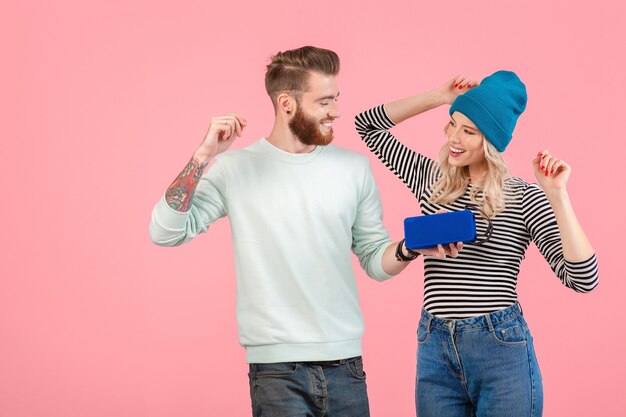 Atractiva pareja joven escuchando música en altavoz inalámbrico vistiendo un traje elegante y fresco sonriendo feliz estado de ánimo positivo posando sobre fondo rosa aislado bailando divirtiéndose