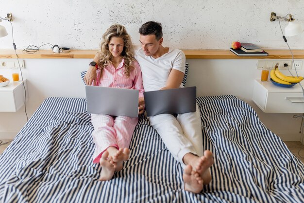 Atractiva pareja joven sentada en la cama por la mañana, hombre y mujer trabajando en la computadora portátil