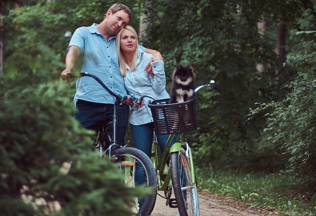 Una atractiva pareja de una mujer rubia y un hombre se abrazan, vestidos con ropa informal en un paseo en bicicleta con su pequeño y lindo spitz en una canasta.