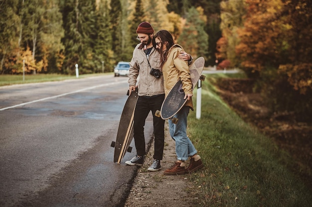 Atractiva pareja romántica camina por la carretera rodeada de árboles de otoño mientras sostiene sus longboards.