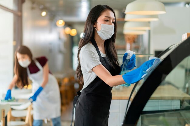 Atractiva y positiva personal asiática joven con delantal uniforme rociando detergente o alcohol en el gabinete de productos de mostrador limpiándolo con un paño suave al desinfectar el café por la mañana seguro y limpio