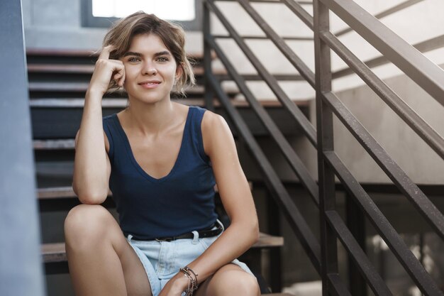 Atractiva rubia europea joven estudiante sentada sola al aire libre escaleras mirar cámara hablando amigo descansando después de la lección universidad caminando campus relajante disfrutar verano cálido clima usar pantalones cortos