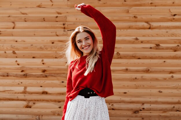 Atractiva rubia en un hermoso suéter rojo pasando un buen rato al aire libre. Mujer joven sonriente posando felizmente en la pared de madera.