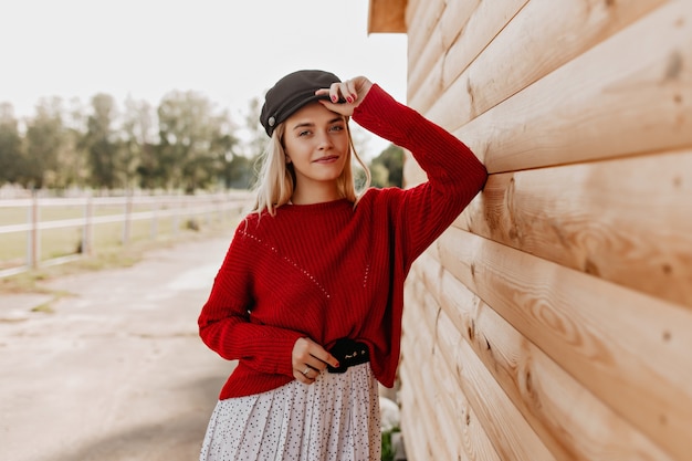 Atractiva rubia de maquillaje natural que mira con ternura. Mujer joven en jersey rojo y elegante sombrero posando cerca de la casa de madera.