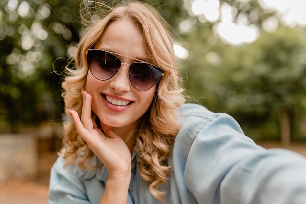 Atractiva rubia sonriente mujer de dientes blancos caminando en el parque en traje de verano tomando foto selfie en teléfono